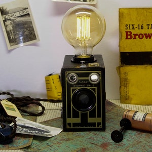 May include: A vintage camera lamp with a black and gold finish. The lamp is made from a repurposed Kodak Brownie Six-16 camera. The lamp has a clear glass bulb with a filament visible. The lamp is sitting on a table with a roll of film and a few other items.