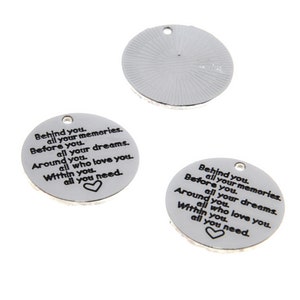 Op de afbeelding: Twee ronde zilveren bedels met de tekst "Behind you, all your memories. Before you, all your dreams. Around you, all who love you. Within you, all you need." en een hartje.