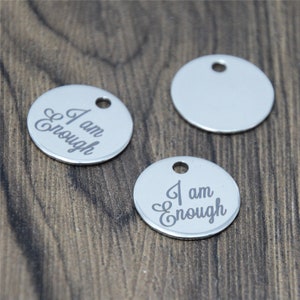May include: Three silver metal round charms with a hole at the top. Two of the charms have the words "I am Enough" engraved on them. The third charm is blank.