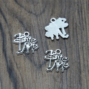 May include: Two silver charm pendants with the words "Bite Me" in a gothic font. The charms are on a wooden surface.