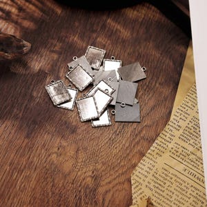 Puede incluir: Una colección de pequeños colgantes cuadrados plateados. Cada colgante tiene una superficie texturizada y un lazo para sujetarlo a una cadena o cordón. Algunos tienen una superficie clara y reflectante, otros un diseño.