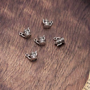 May include: Several small, silver-colored teapot charms are scattered on a wooden surface. Each charm has a detailed design, including a handle, spout, and decorative elements. The charms are ideal for jewelry making or crafting projects.