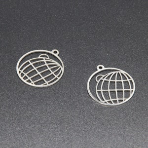 Op de afbeelding: Twee zilveren metalen bedels in de vorm van een globe met een cirkel eromheen. De globe is omlijnd en heeft een rasterpatroon.
