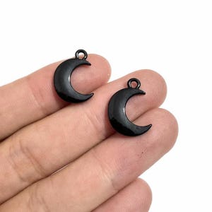 May include: Two small, glossy black crescent moon charms, each featuring a circular jump ring at the top for attachment. The charms have a smooth, reflective surface and a curved, celestial shape, presented as individual jewelry components.