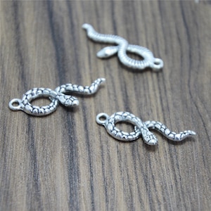 Puede incluir: Tres colgantes plateados con forma de serpiente con un diseño detallado y texturizado. Cada colgante presenta una forma de serpiente enrollada con un lazo en cada extremo para sujetar. Se muestran sobre una superficie de madera.