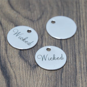 May include: Three silver-coloured, circular charms with a hole at the top. Two charms have the word "Wicked" engraved in a cursive font. The charms are on a brown wooden surface.