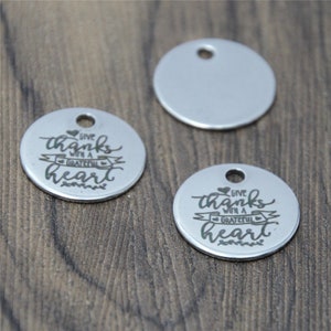 May include: Two silver metal round charms with the text "Give thanks with a grateful heart" engraved on them. The charms have a small hole at the top for attaching to a necklace or bracelet.