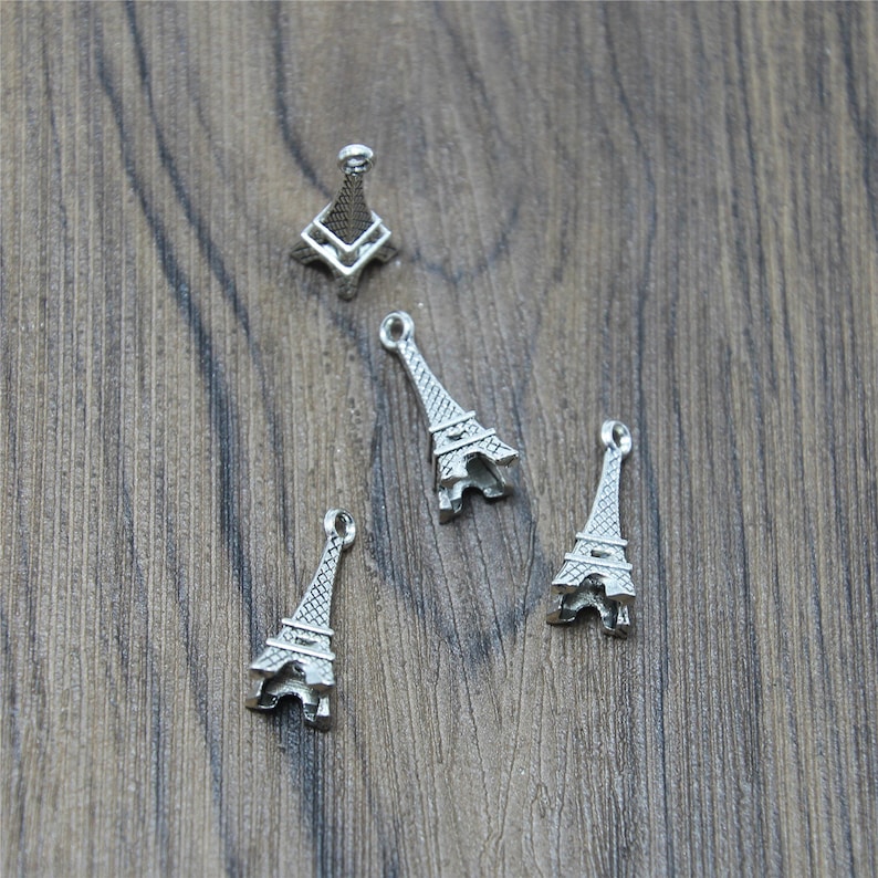May include: Four silver Eiffel Tower charms, each with a detailed design, are arranged on a wooden surface.