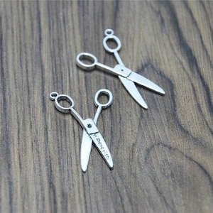 Puede incluir: Dos abalorios de tijeras plateadas con un lazo para colgar. El abalorio más pequeño tiene el texto "Misengyi.sh" grabado en una hoja.