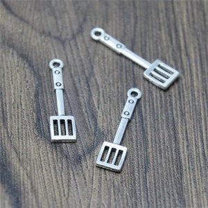 May include: Three silver-colored metal spatula charms with a slotted rectangular head and a handle with a loop at the end. The charms are scattered on a wooden surface.