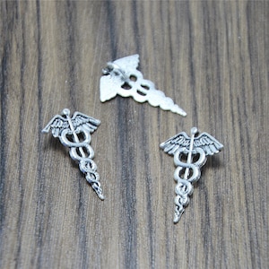 Puede incluir: Tres amuletos de caduceo médico de plata con una serpiente y alas. Los amuletos están sobre una superficie de madera.