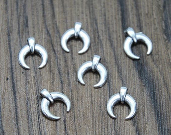 May include: Six small, silver-colored crescent moon charms. Each charm has a curved horn shape with a small rectangular bar at the top. The charms are scattered on a wooden surface.