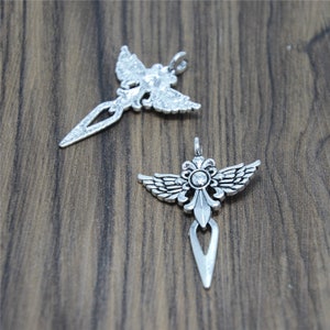 May include: Two silver-toned pendants with an angel wing design. One pendant features a clear crystal in the center. The pendants have a decorative, ornate design and are displayed on a wooden surface.