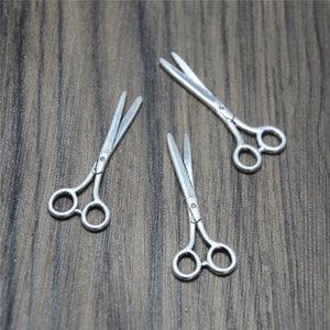 May include: Three miniature silver-toned scissors with pointed tips and rounded handles. The scissors are arranged in a triangular pattern on a wooden surface.