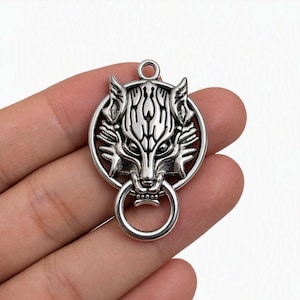 May include: A silver-toned metal pendant featuring a stylised wolf head design enclosed within a circular frame. The wolf head has intricate engraved details and holds a small, smooth circular ring in its mouth. A small loop is at the top.
