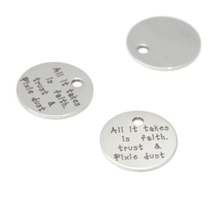 Może przedstawiać: Dwa srebrne, okrągłe breloki z otworem na górze. Breloki mają wygrawerowany tekst "All it takes is faith. trust & pixie dust".