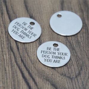 May include: Three silver-colored, circular dog tags with a hole at the top. Two tags have the engraved text: "BE THE PERSON YOUR DOG THINKS YOU ARE." The tags are on a wooden surface.