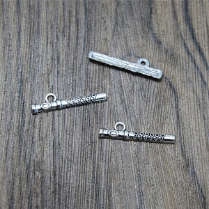 Könnte beinhalten: Drei silberne Metall-Anhänger in Form einer Flöte. Die Anhänger haben oben eine Schlaufe zum Anbringen an einer Halskette oder einem Armband.