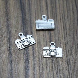 May include: Three silver-toned camera charms. Each charm features a detailed camera design with a lens and a small loop for attaching to a necklace or bracelet.