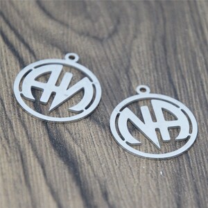 May include: Two silver metal charms with the letters "NA" and "AM" in a circle design.