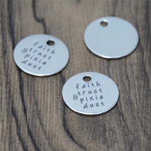 Puede incluir: Dos dijes redondos de metal plateado con las palabras "faith trust pixie dust" grabadas en ellos.
