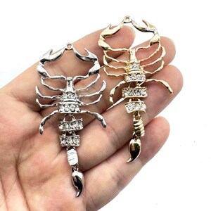 Op de afbeelding: Twee schorpioenhangers, één zilverkleurig en één goudkleurig, worden getoond. Elke hanger heeft een gedetailleerd schorpioenontwerp met strass accenten. De hangers worden in een hand gehouden tegen een witte achtergrond.