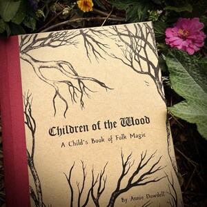 Op de afbeelding: Een boek getiteld "Children of the Wood: A Child's Book of Folk Magic" door Annie Dowdell. De omslag heeft een bruine achtergrond met zwarte inkt illustraties van bomen en takken.