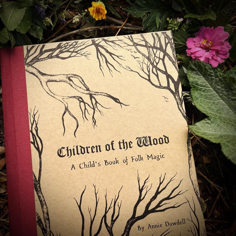 Op de afbeelding: Een boek getiteld "Children of the Wood: A Child's Book of Folk Magic" door Annie Dowdell. De omslag heeft een bruine achtergrond met zwarte inkt illustraties van bomen en takken.