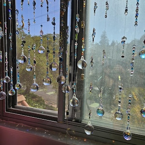 May include: A window adorned with multiple strands of decorative crystal beads and orbs. The strands hang in front of the window, reflecting sunlight and creating a shimmering effect. The beads are various colors and shapes, adding visual interest.