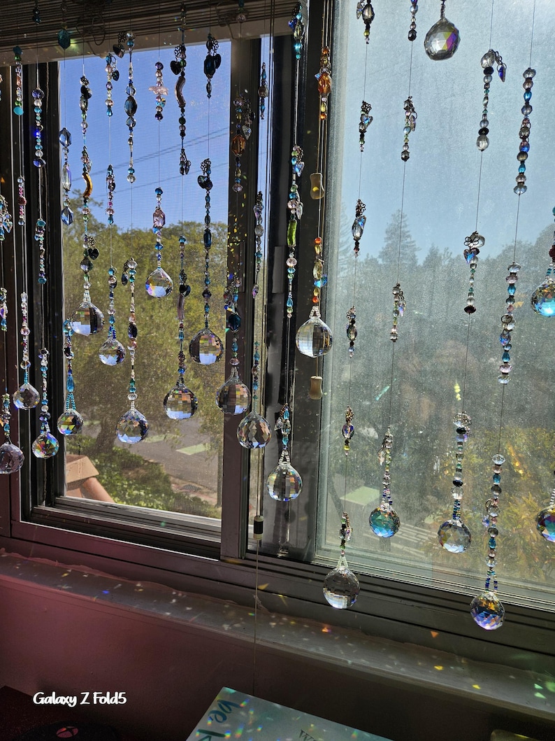 May include: A window adorned with multiple strands of decorative crystal beads and orbs. The strands hang in front of the window, reflecting sunlight and creating a shimmering effect. The beads are various colors and shapes, adding visual interest.
