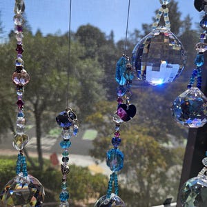 May include: Several crystal suncatchers hang in front of a window. Each suncatcher features a large, faceted crystal ball, with smaller beads in shades of blue, purple, and clear. The suncatchers are suspended by thin, silver-colored wires.
