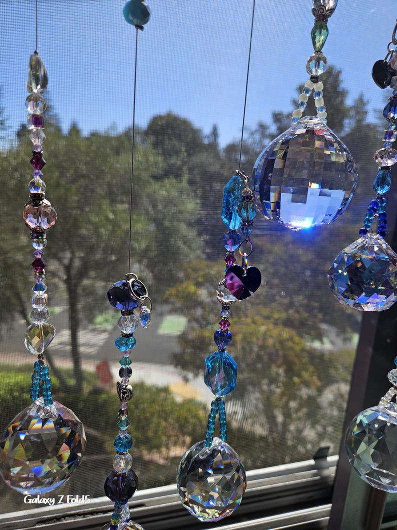 May include: Several crystal suncatchers hang in front of a window. Each suncatcher features a large, faceted crystal ball, with smaller beads in shades of blue, purple, and clear. The suncatchers are suspended by thin, silver-colored wires.
