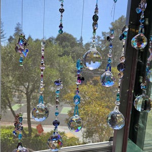 May include: Decorative crystal suncatchers hanging in front of a window. The suncatchers feature faceted crystal balls, beads, and heart-shaped accents in shades of blue, purple, and clear. The window frame is visible, with a view of trees and sky.