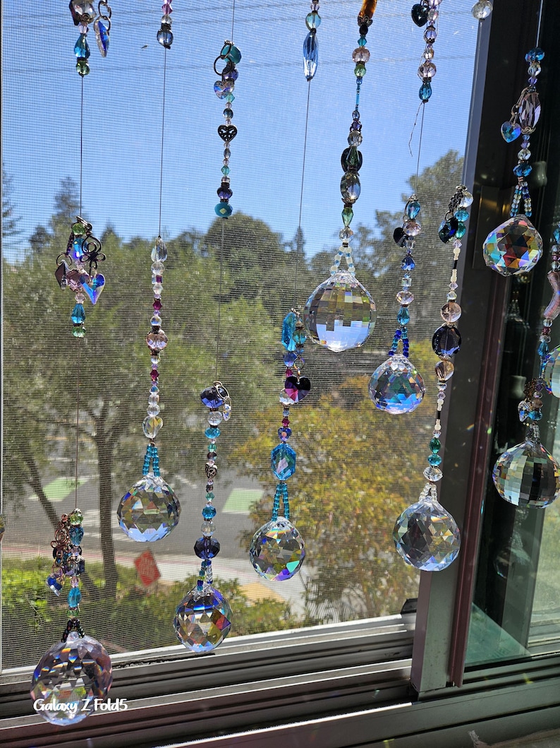 May include: Decorative crystal suncatchers hanging in front of a window. The suncatchers feature faceted crystal balls, beads, and heart-shaped accents in shades of blue, purple, and clear. The window frame is visible, with a view of trees and sky.