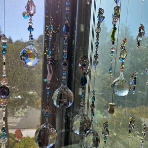 May include: A collection of hanging crystal suncatchers with various shapes and sizes. The suncatchers feature clear, blue, and purple crystals, with some having decorative metal accents. They are suspended in front of a window, catching the sunlight.