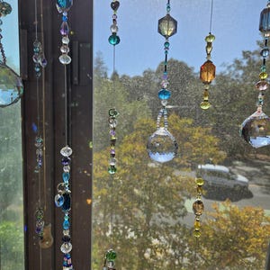 May include: A collection of hanging crystal suncatchers with various shapes and colors, including clear, blue, purple, and amber. The suncatchers are suspended in front of a window, creating a shimmering display. The image is taken in natural light.
