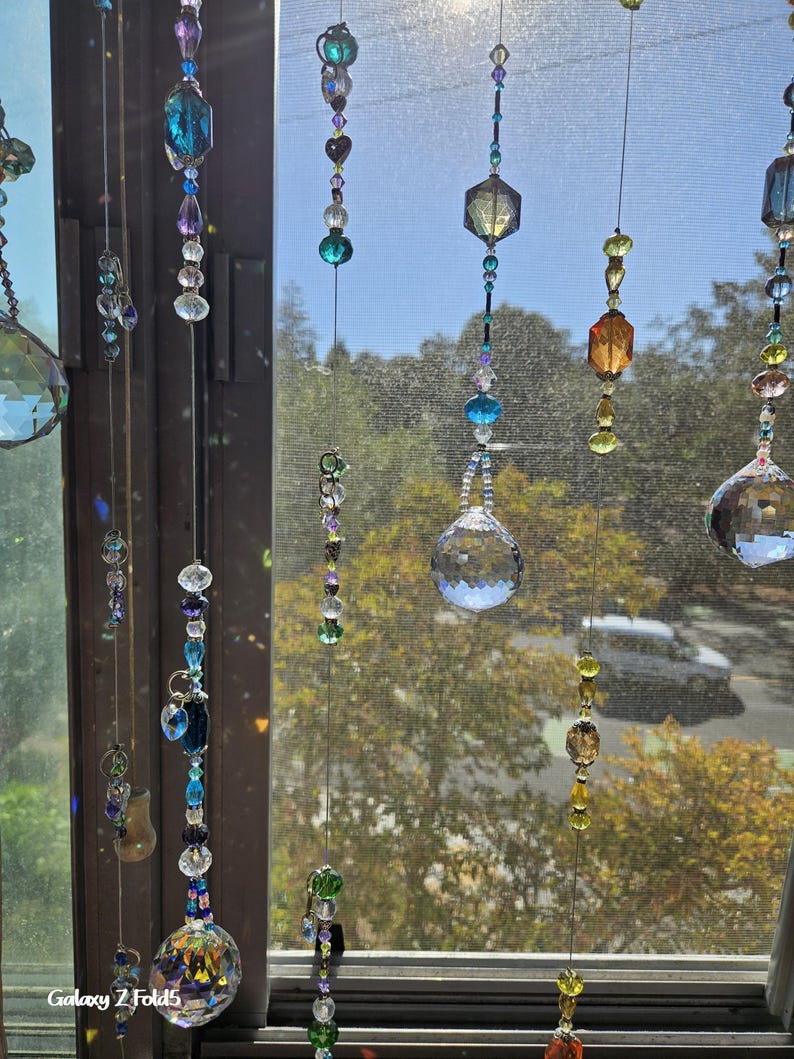 May include: A collection of hanging crystal suncatchers with various shapes and colors, including clear, blue, purple, and amber. The suncatchers are suspended in front of a window, creating a shimmering display. The image is taken in natural light.