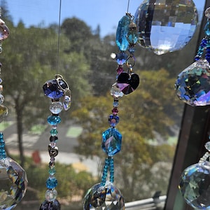 May include: Close-up of hanging crystal suncatchers with various shapes and sizes of clear and blue crystals. The suncatchers are strung together with beads and hang in front of a window with a blurred outdoor background.