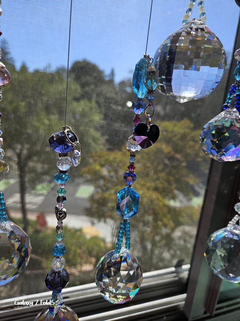 May include: Close-up of hanging crystal suncatchers with various shapes and sizes of clear and blue crystals. The suncatchers are strung together with beads and hang in front of a window with a blurred outdoor background.