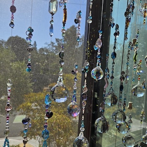 May include: A collection of hanging crystal suncatchers with various shapes and sizes. The suncatchers are strung together with beads in shades of blue, purple, and clear. The background shows a window with trees and a bright sky.