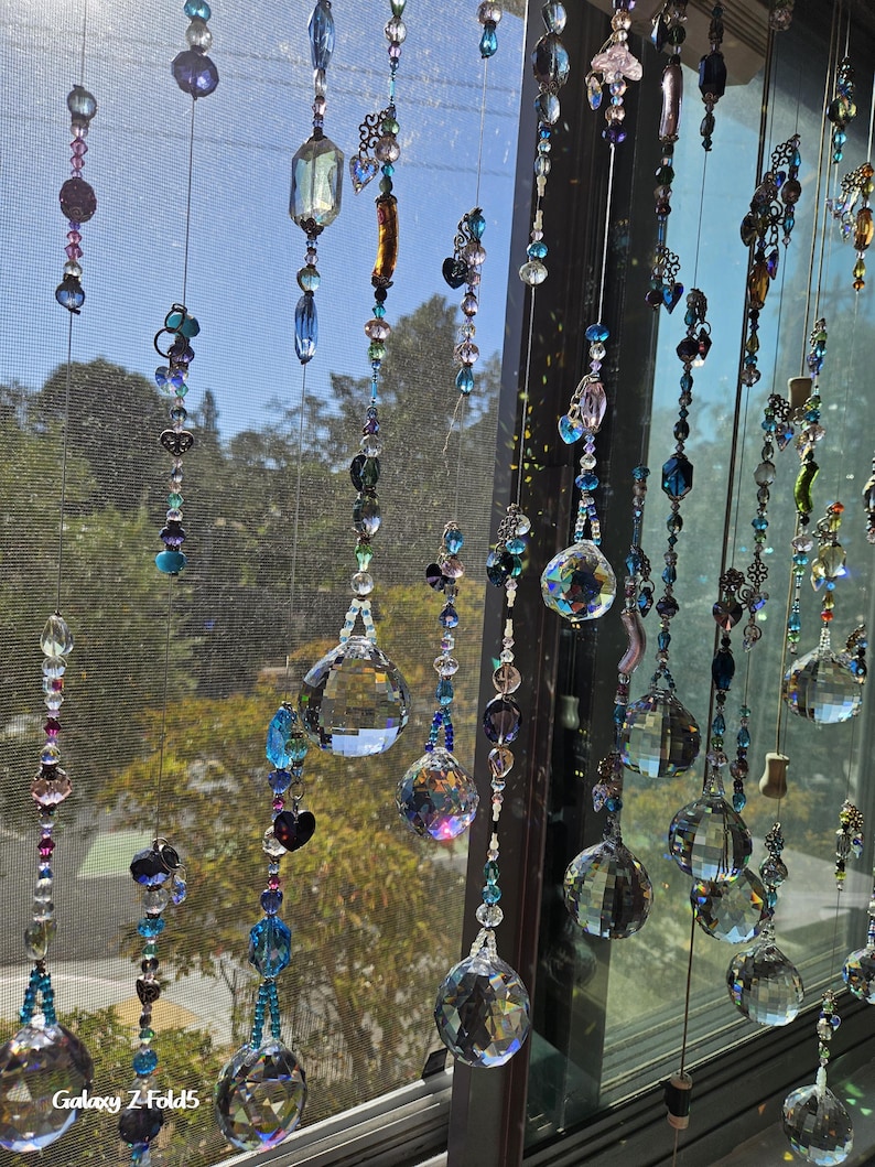 May include: A collection of hanging crystal suncatchers with various shapes and sizes. The suncatchers are strung together with beads in shades of blue, purple, and clear. The background shows a window with trees and a bright sky.