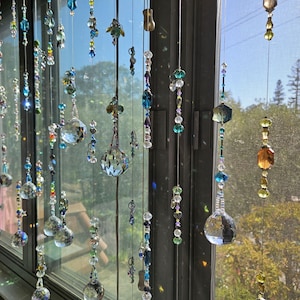 May include: A window display featuring multiple strands of decorative crystal beads in various shapes and sizes. The beads are in shades of blue, green, and clear, with some iridescent elements. The strands hang in front of a window, creating a shimmering effect.