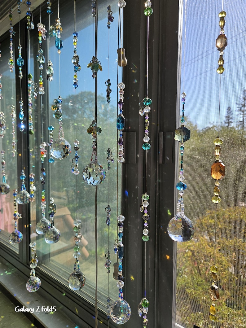 May include: A window display featuring multiple strands of decorative crystal beads in various shapes and sizes. The beads are in shades of blue, green, and clear, with some iridescent elements. The strands hang in front of a window, creating a shimmering effect.