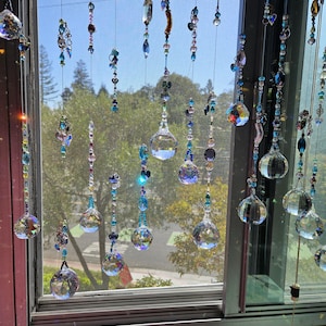May include: Decorative crystal beaded window hangings. The strands feature clear crystal spheres and various blue and clear beads. The window is open, with a view of trees and a blue sky.