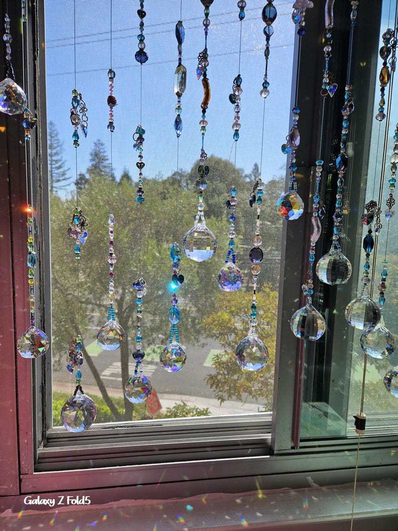 May include: Decorative crystal beaded window hangings. The strands feature clear crystal spheres and various blue and clear beads. The window is open, with a view of trees and a blue sky.