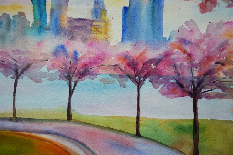 以下が含まれることがあります： 水彩画で描かれた街並みの風景画。前景にはピンクの桜の木が描かれている。木はピンク、パープル、ホワイトの色合いで描かれており、背景には淡いブルーの空が広がっている。
