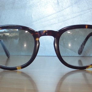 Könnte beinhalten: Brille mit Schildpattmuster, abgerundeten Gläsern und Schlüssellochsteg. Die Gläser haben einen leichten Farbverlauf und die Bügel sind an den äußeren Ecken des Rahmens mit kleinen, silberfarbenen Metalldetails verziert.