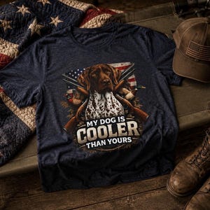 Puede incluir: Camiseta azul marino con un gráfico de un perro de caza, armas y una bandera estadounidense. El texto "MY DOG IS COOLER THAN YOURS" está impreso en la camiseta. Se ve una gorra de béisbol marrón con un parche de la bandera estadounidense.