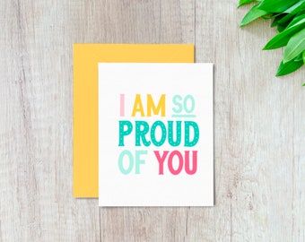Könnte beinhalten: Eine weiße Grußkarte mit dem Text "I AM SO PROUD OF YOU" in bunten Buchstaben. Dazu ein gelber Umschlag. Der Hintergrund ist eine helle Holzoberfläche.
