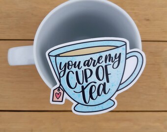 Könnte beinhalten: Eine blaue Teetasse mit dem Text "You are my cup of tea" und einem rosa Teebeutel mit einem Herz.